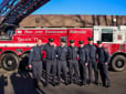 A group of new fire academy graduates in dress uniform smile in front of bright red Truck 71. The truck’s ladder extends over their heads.