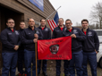 A group of seven fire recruits holds a bright-red class flag outside the Fire Training Center.